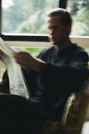 Man seated in an armchair holding and reading a newspaper indoors near a bright window