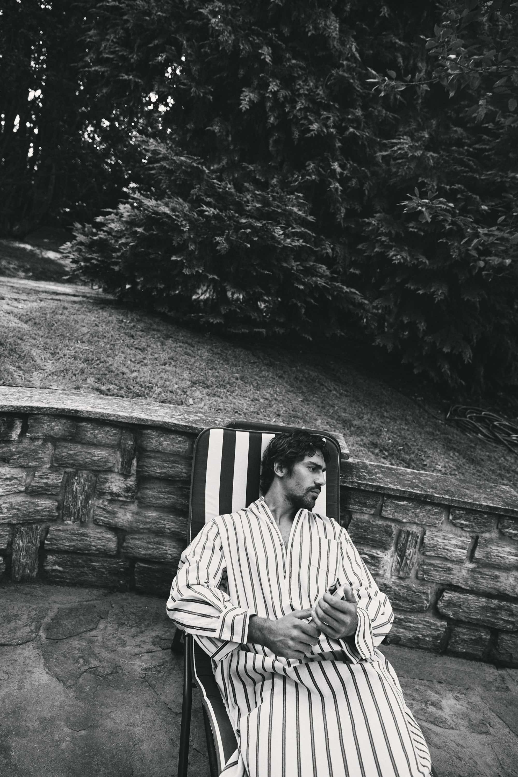 Monochrome portrait of a man in a striped caftan seated on a lounger outdoors against a stone wall and lush foliage.