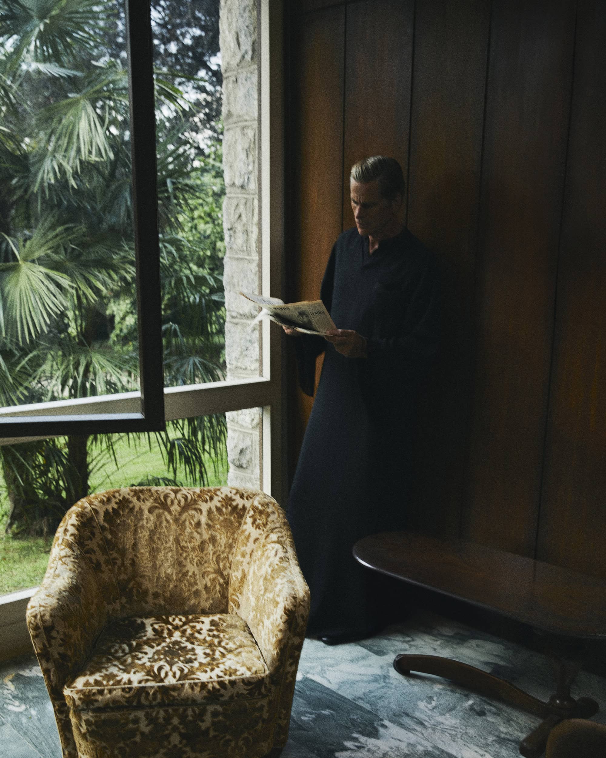 Man in dark robe reading newspaper by large window overlooking palm trees inside a retro interior