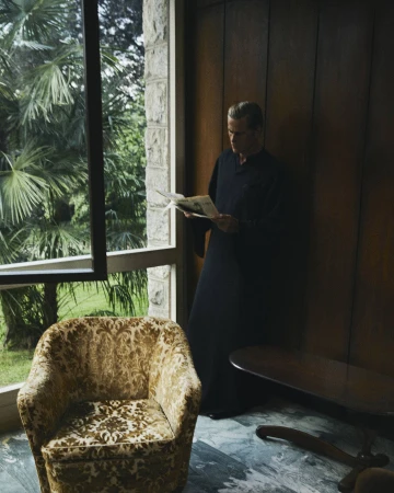 Man in dark robe reading newspaper by large window overlooking palm trees inside a retro interior