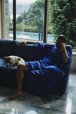 Man in a royal blue kaftan seated on a dark blue sofa next to a small dog with large windows showing lush greenery outside.