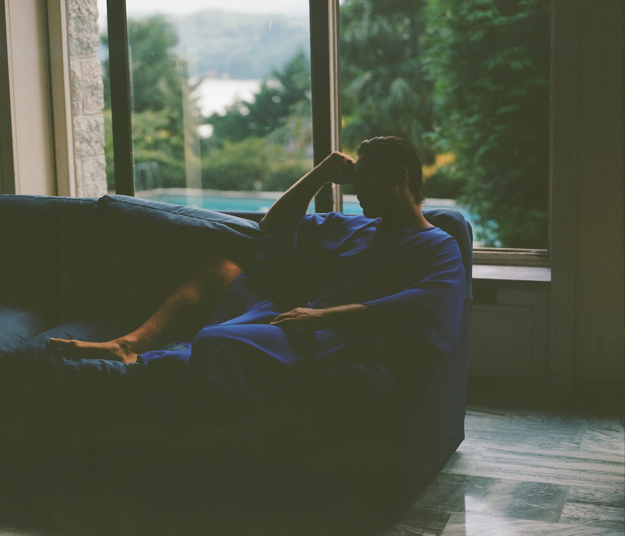 Man relaxing on a blue sofa in front of large windows overlooking a scenic landscape for Taller Marmo campaign.