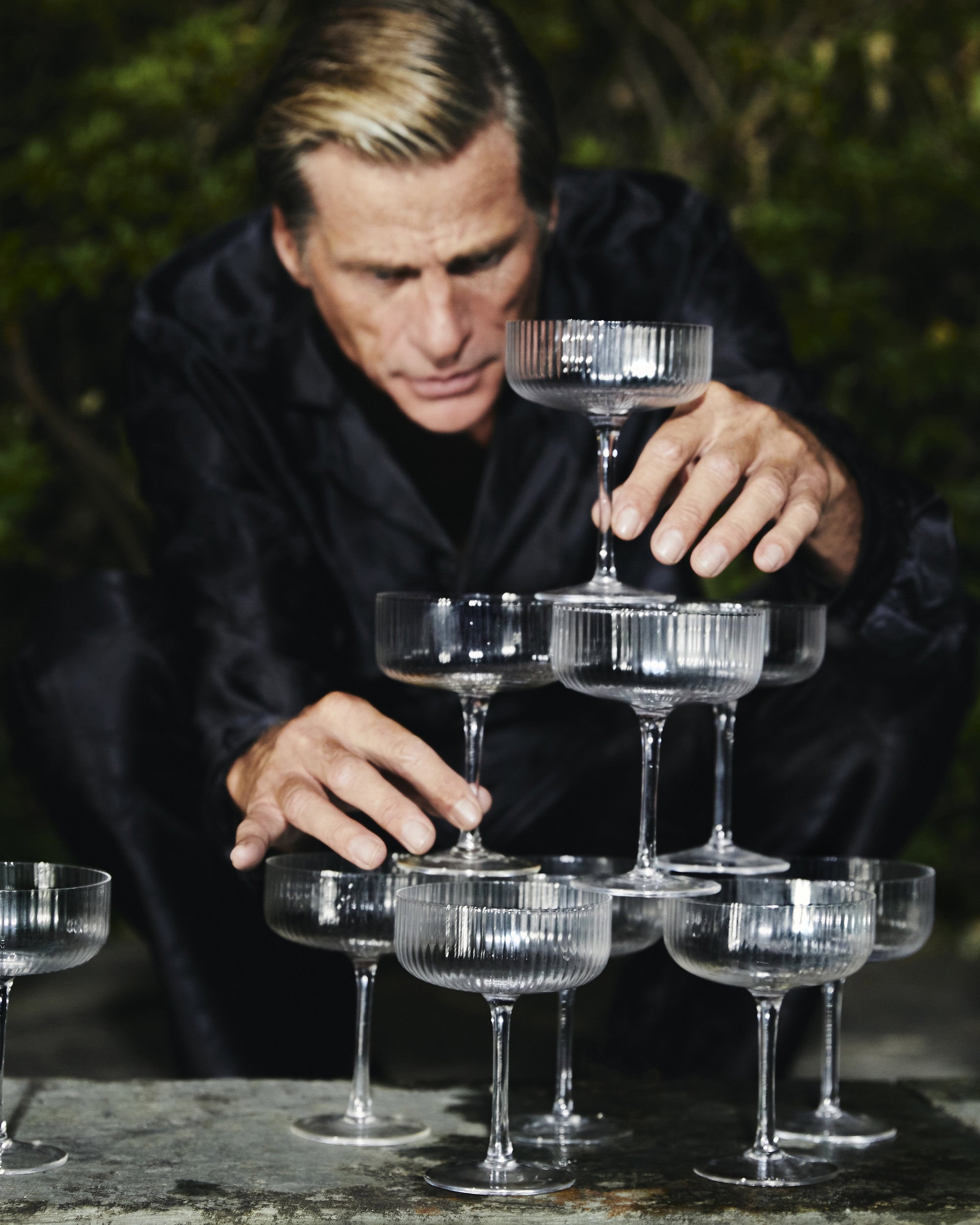 A man wearing a black shirt carefully builds a pyramid tower of ribbed crystal champagne glasses outdoors.