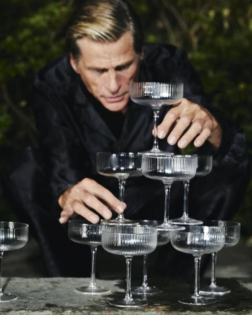 A man wearing a black shirt carefully builds a pyramid tower of ribbed crystal champagne glasses outdoors.