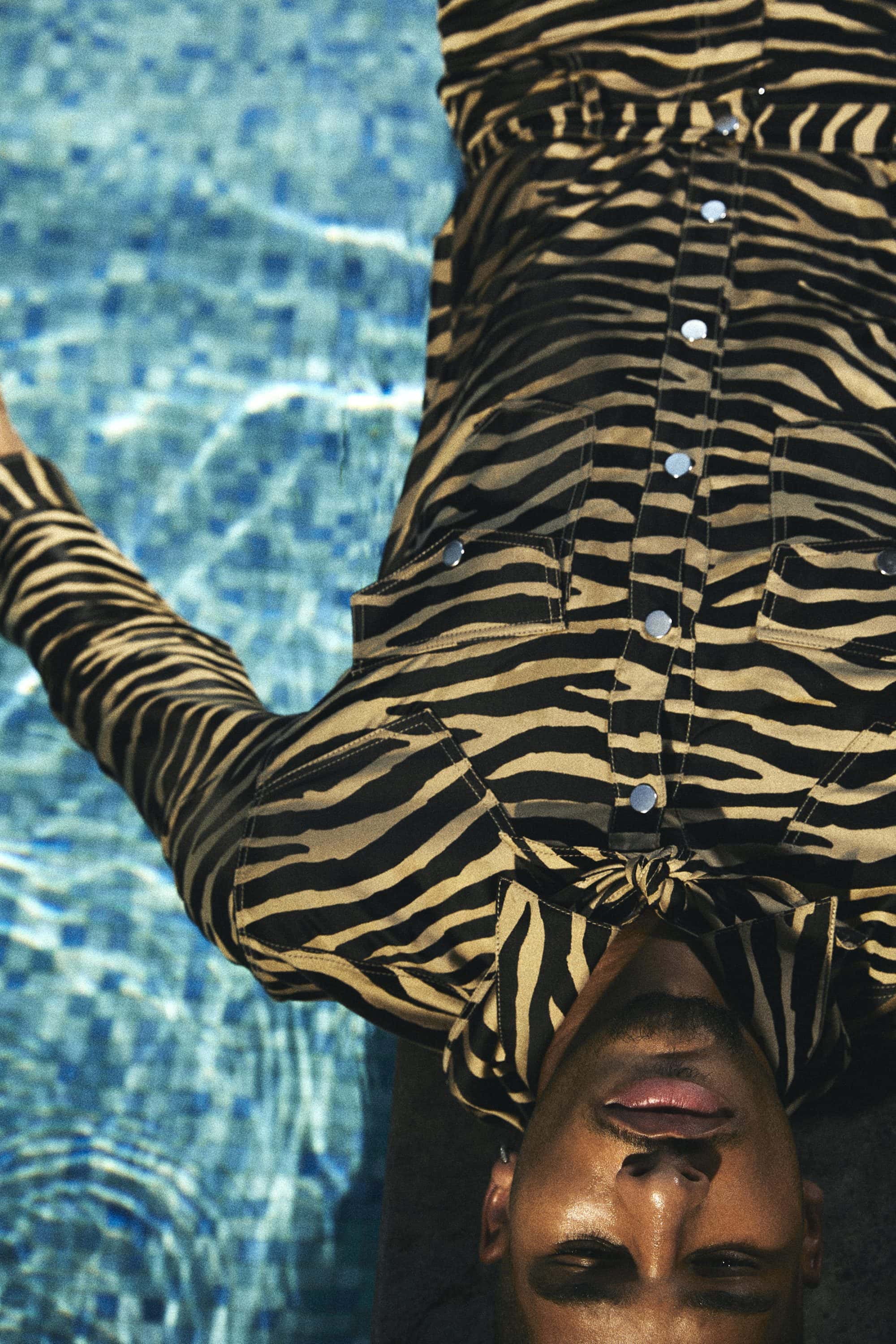 A man wearing a zebra-print long-sleeve shirt lies near a blue tiled swimming pool.