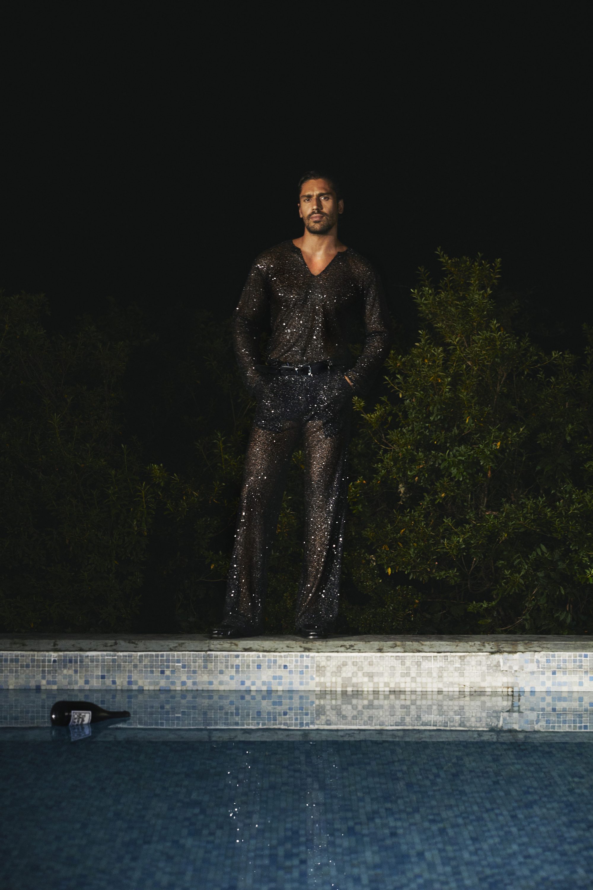 Man wearing a dark sequin top and trousers standing by a tiled swimming pool at night.