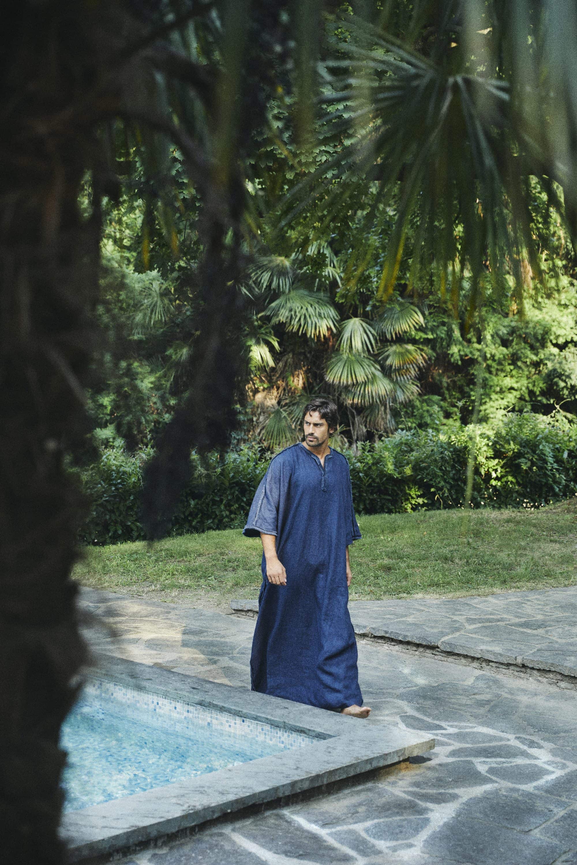 Man wearing a long blue kaftan walking near a stone pool in a lush garden setting by Francesco Scotti