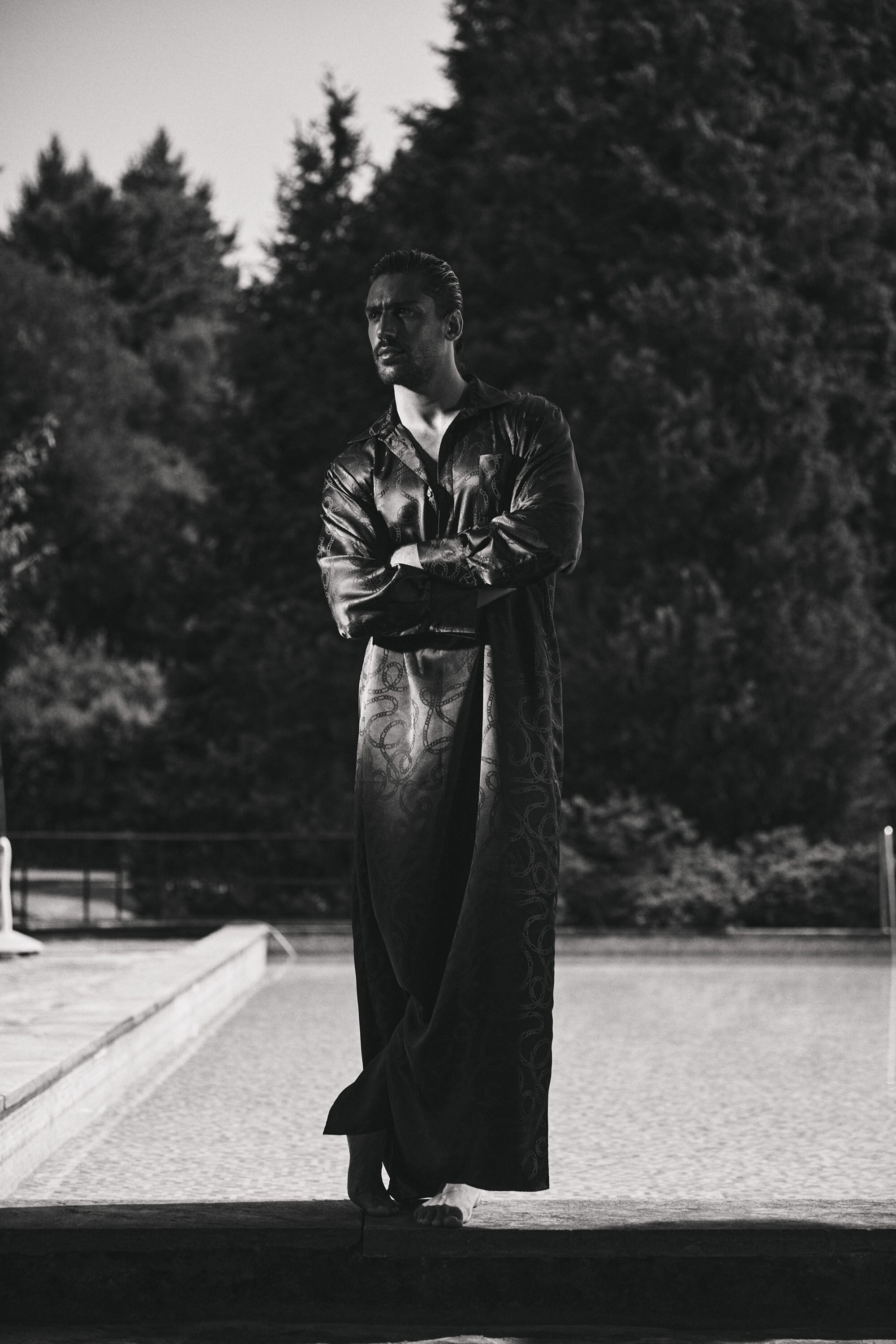 Man standing outdoors by a swimming pool wearing a long dark patterned kaftan with arms crossed.