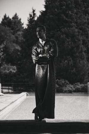 Man standing outdoors by a swimming pool wearing a long dark patterned kaftan with arms crossed.