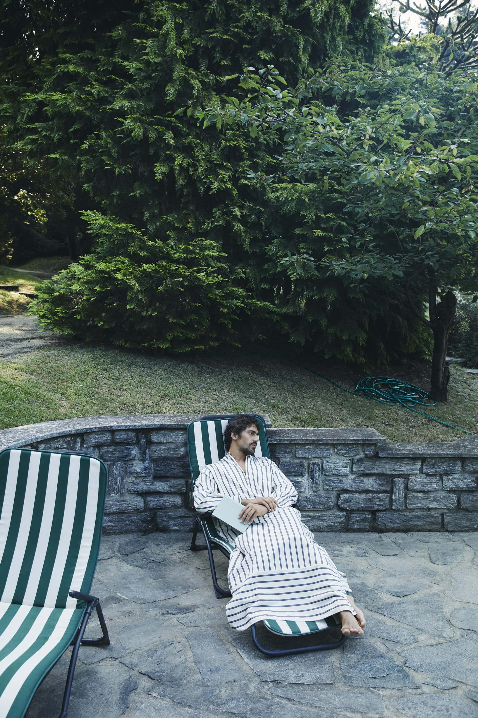 Man resting on a striped lounge chair wearing a striped robe in a lush outdoor garden setting.