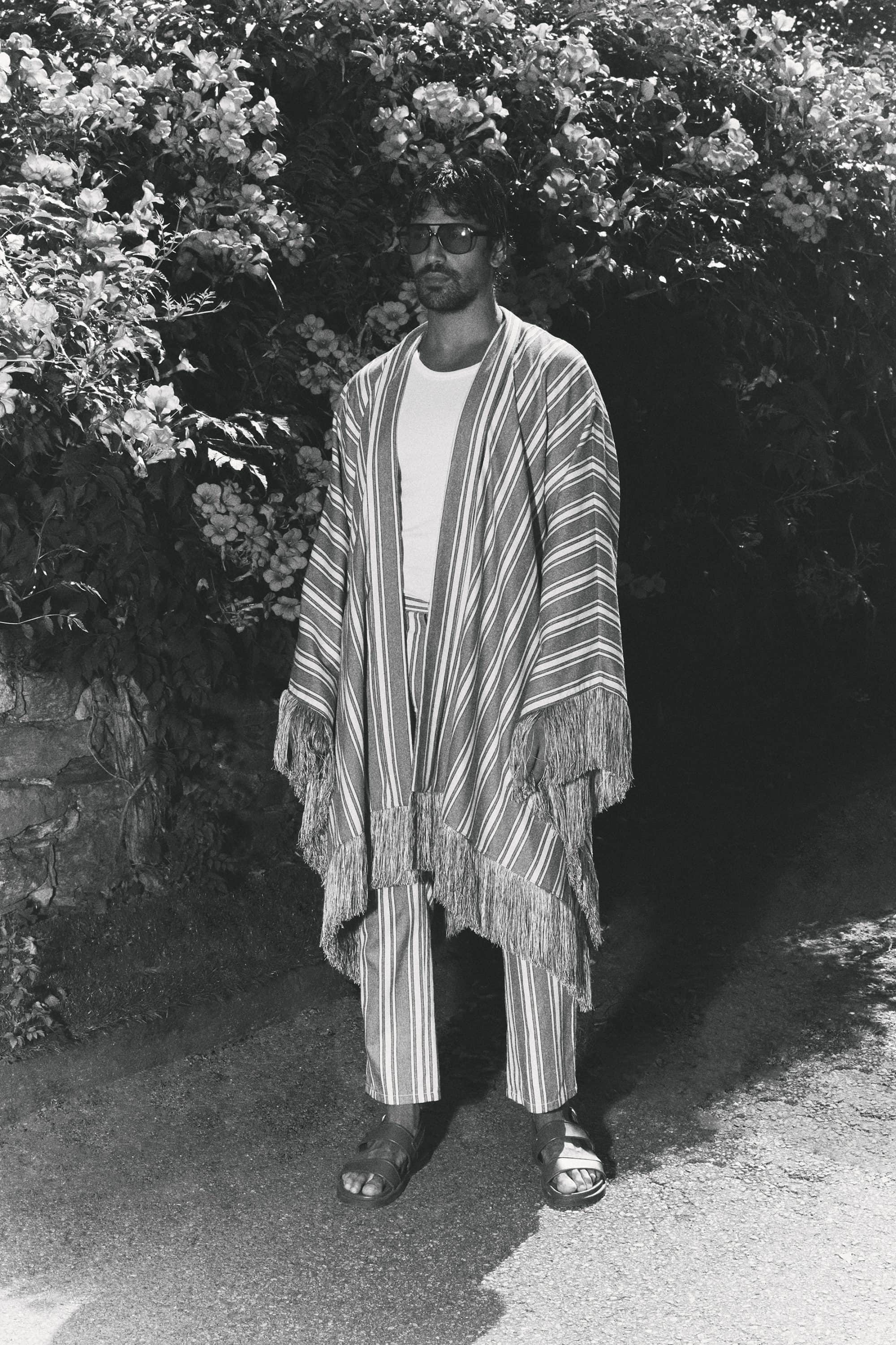 Man wearing a patterned striped fringed robe and trousers standing on an outdoor path in front of leafy foliage.