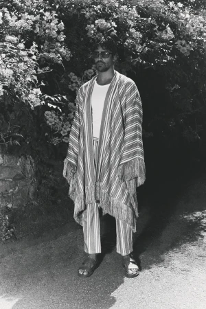 Man wearing a patterned striped fringed robe and trousers standing on an outdoor path in front of leafy foliage.