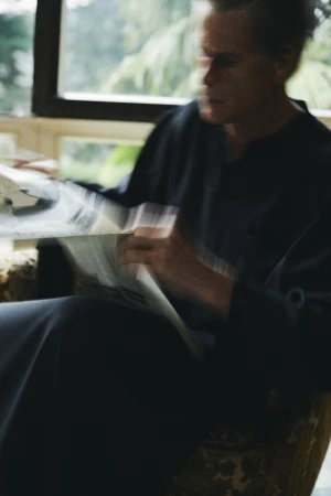 Man wearing a dark shirt seated indoors, reading a newspaper with motion blur against a window.