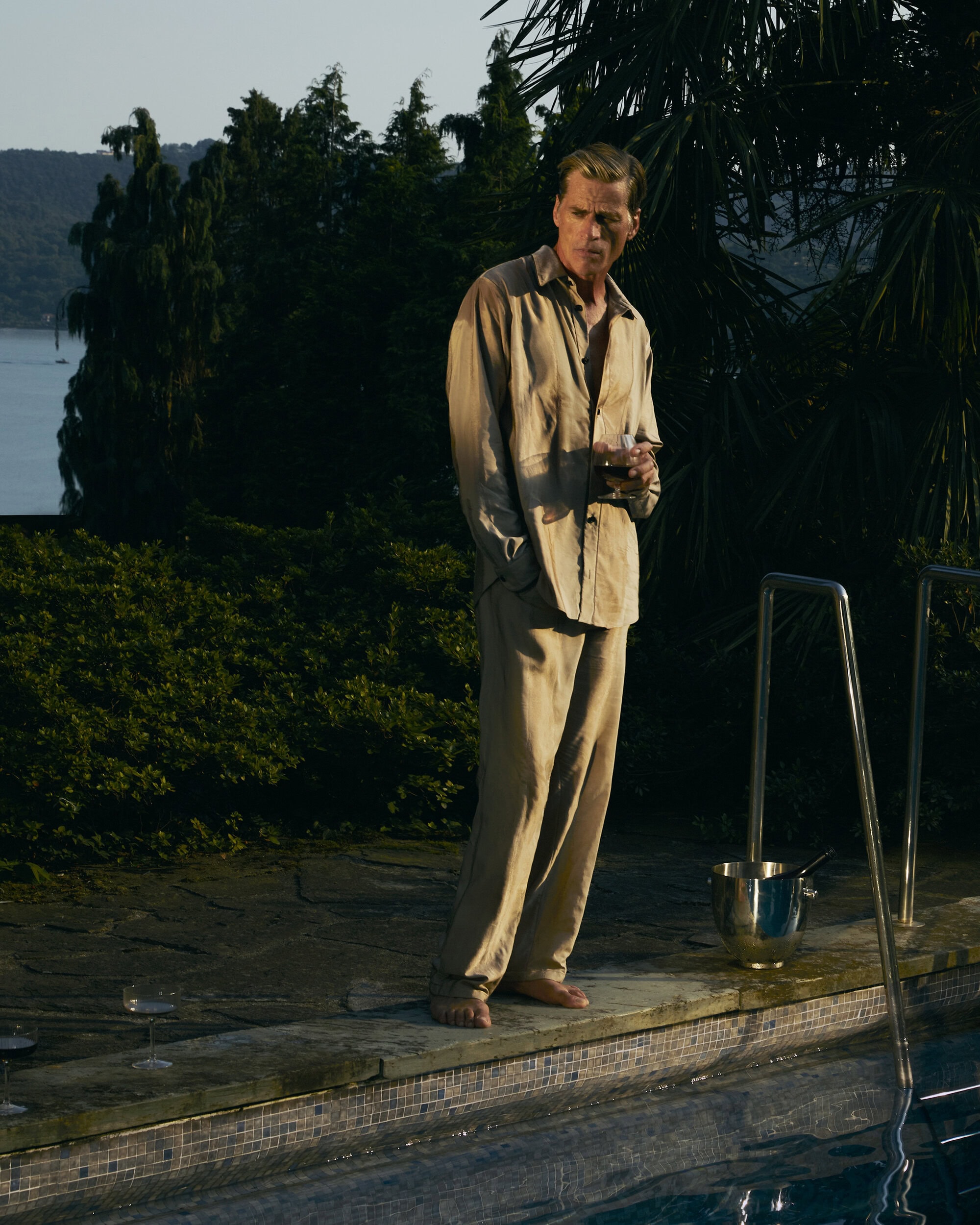Man standing barefoot by a swimming pool at dusk wearing a loose beige shirt and matching trousers.