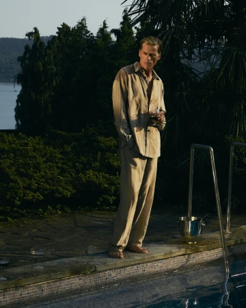 Man standing barefoot by a swimming pool at dusk wearing a loose beige shirt and matching trousers.