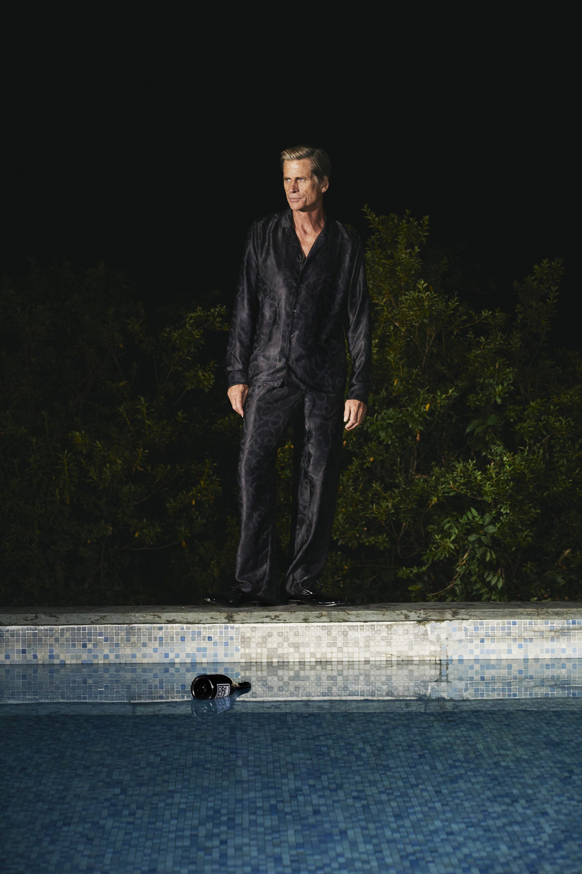Model Mark Vanderloo standing by a tiled swimming pool wearing a dark patterned silk pajama set for Taller Marmo.