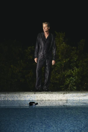 Model Mark Vanderloo standing by a tiled swimming pool wearing a dark patterned silk pajama set for Taller Marmo.