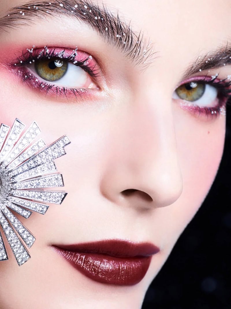 Close-up portrait of model Vittoria Ceretti wearing shimmering pink eye makeup and deep burgundy lipstick with a jeweled brooch.