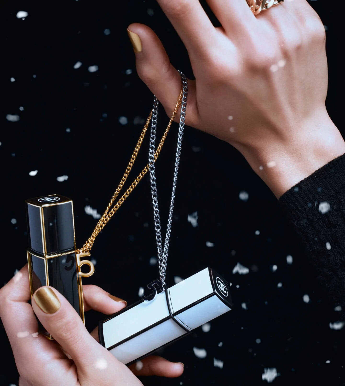 Close-up of hands holding black and white perfume bottles with chains against a dark snowy background.