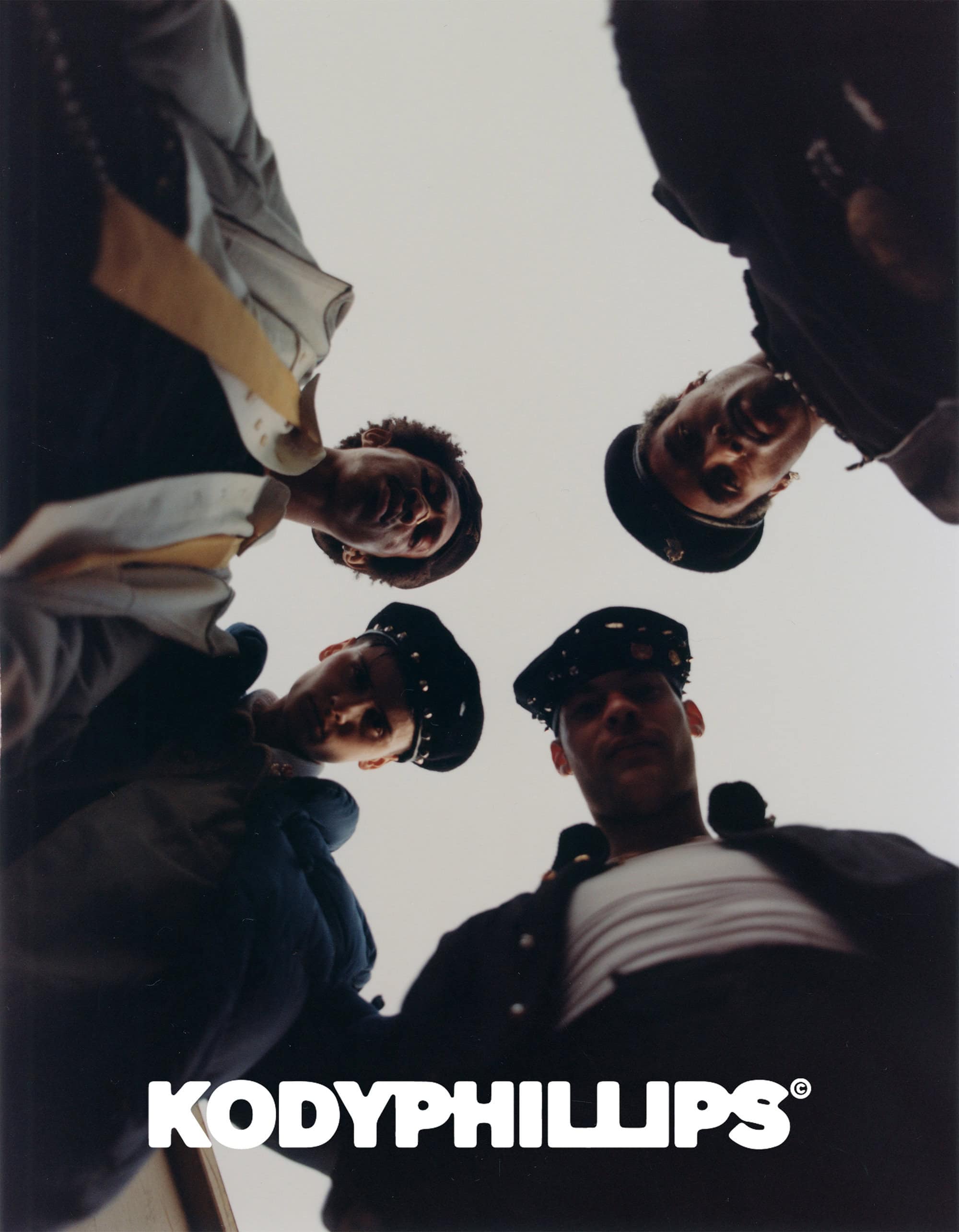 Low angle group shot of four young men looking down wearing dark jackets and caps against a light sky background