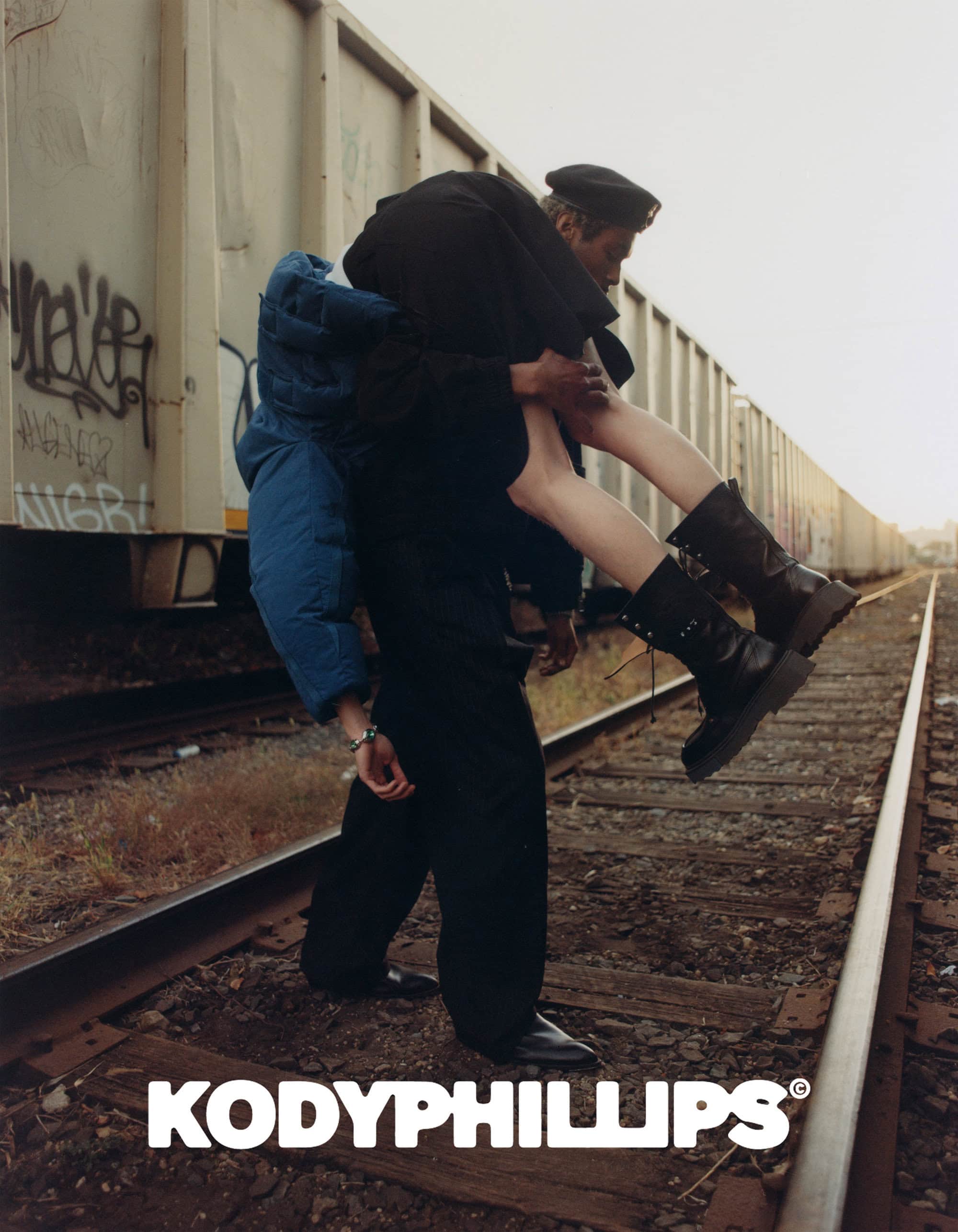 Man carrying a woman over his shoulder while standing on train tracks next to a freight car during Kody Phillips Fall 2024 campaign