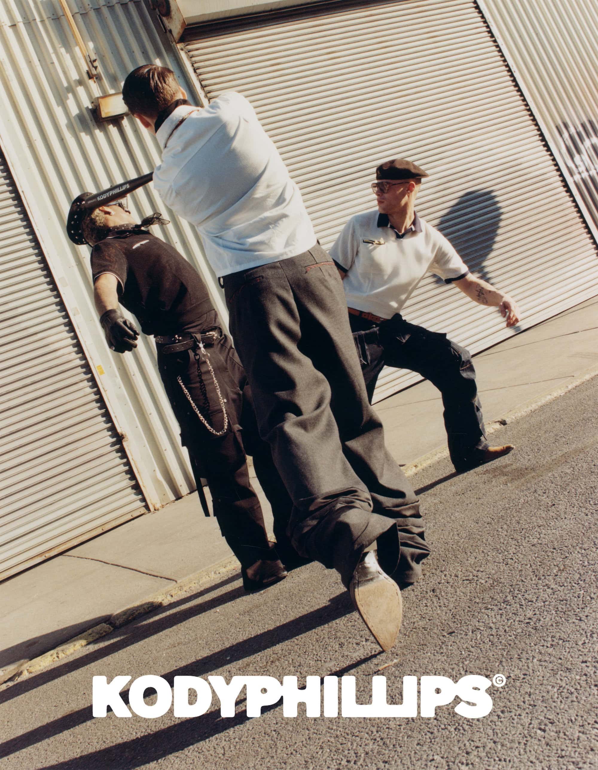 Three men in styled clothing interacting outside corrugated metal building with asphalt ground in the foreground