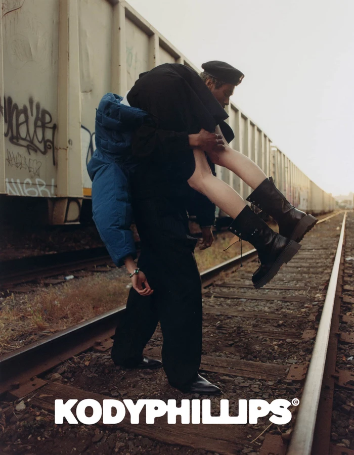 Man carrying a woman over his shoulder while standing on train tracks next to a freight car during Kody Phillips Fall 2024 campaign