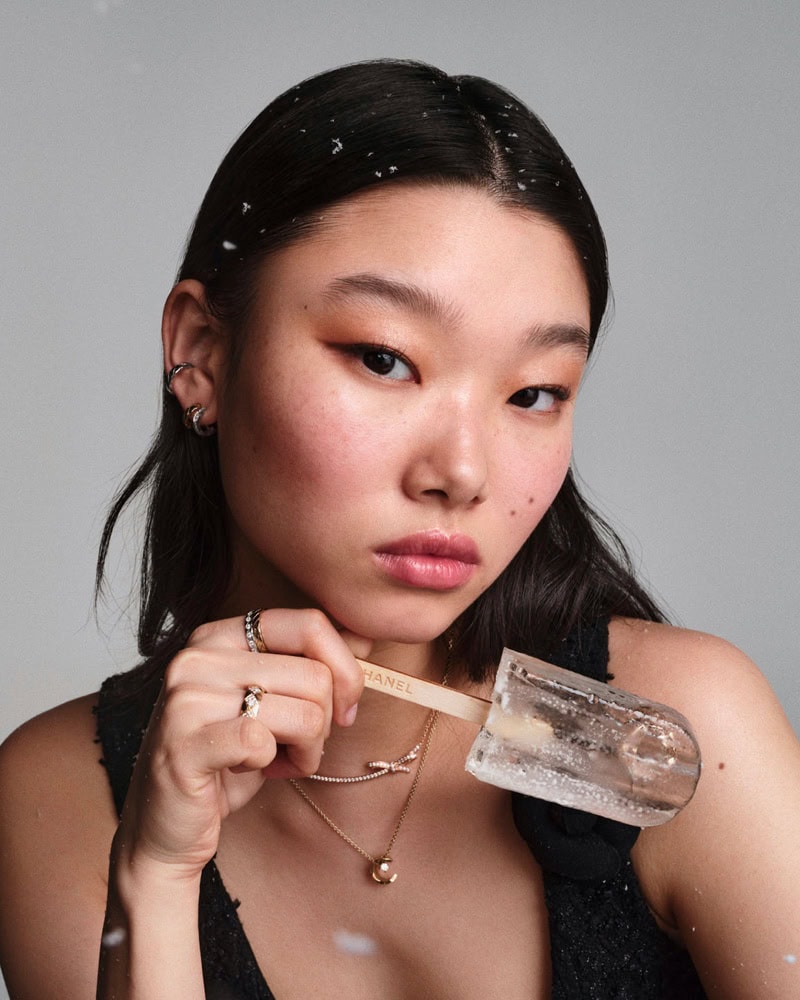 Model holding clear ice popsicle with Chanel branding, wearing fine jewelry and dark top against a neutral background.