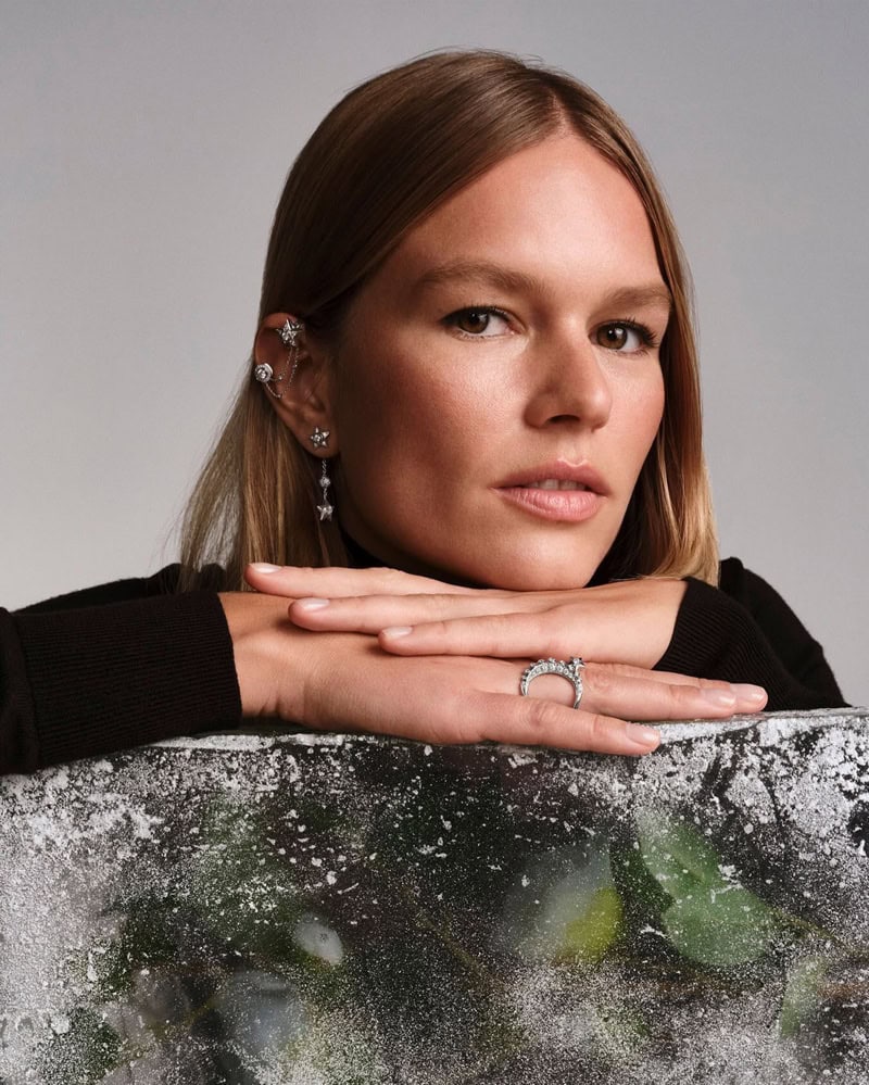 Model Anna Ewers wearing Chanel fine jewelry, resting her chin on folded hands against a neutral grey studio background.