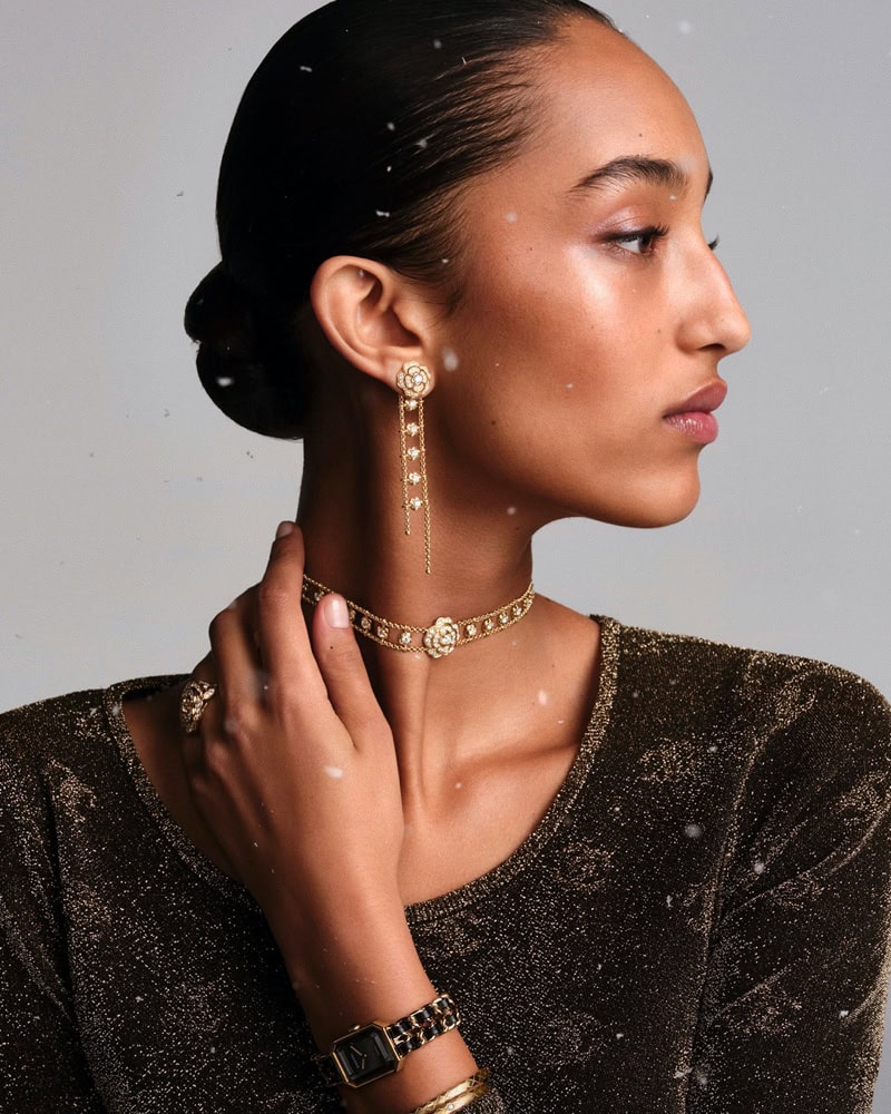 Close-up portrait of a model wearing Chanel fine jewelry including drop earrings, a choker, rings, and a watch against a grey background.