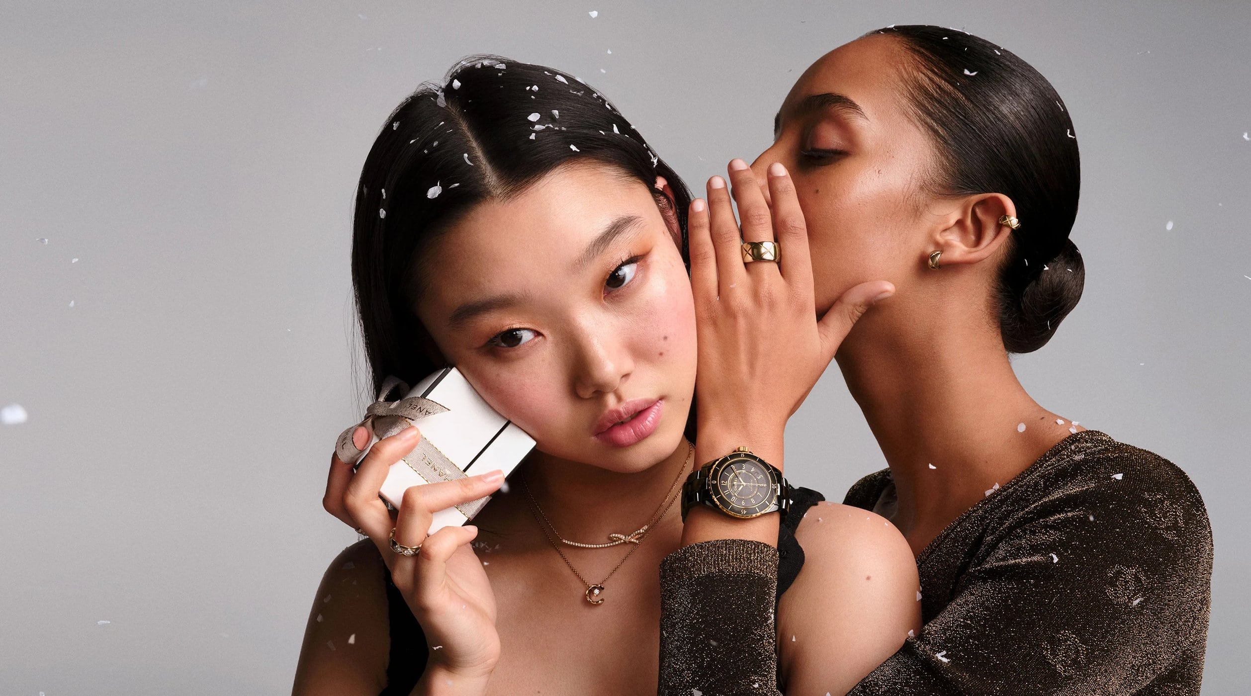 Two models wearing fine jewelry and watches in a Chanel holiday campaign with falling snow effect against a grey background.