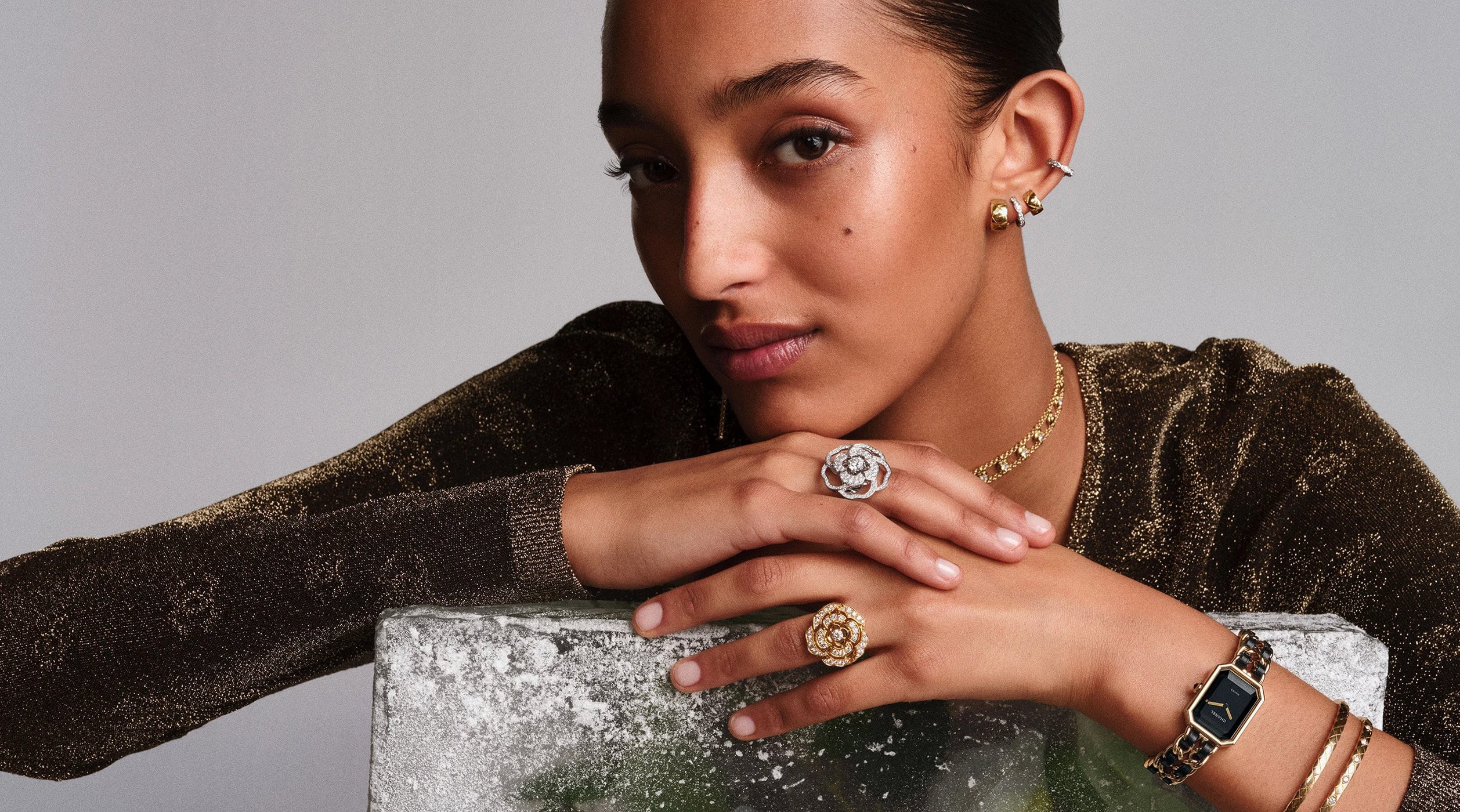 Close-up portrait of a model resting chin on hands wearing fine jewelry and gold wristwatch against grey background