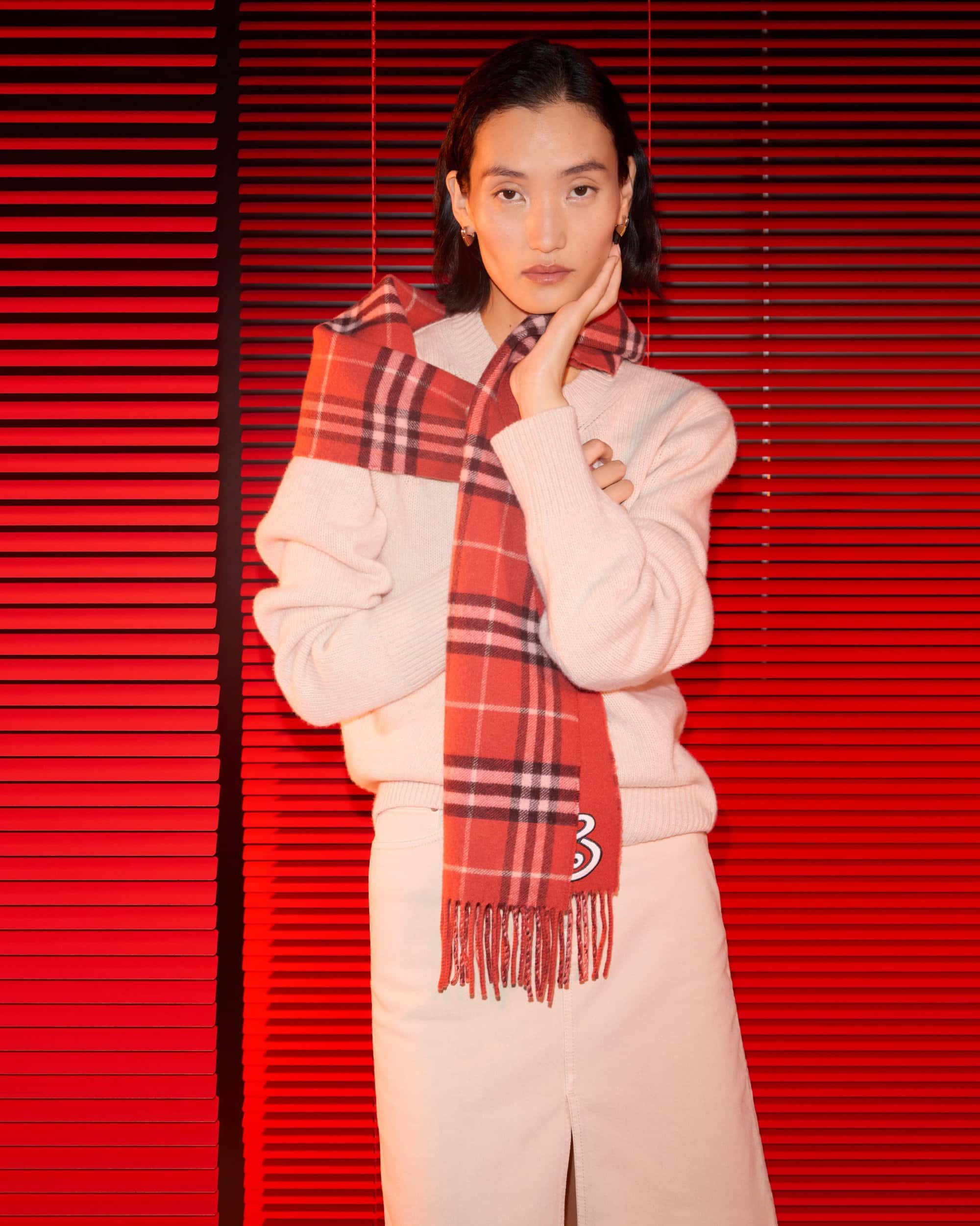 Model wearing a cream knit sweater and plaid scarf against vibrant red horizontal blinds for Burberry Year of the Snake campaign