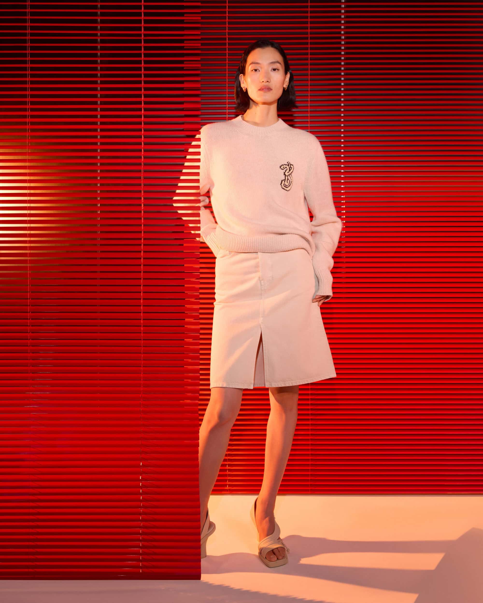 Model in cream knit sweater and skirt standing against bright red slatted blinds for Burberry Year of the Snake campaign