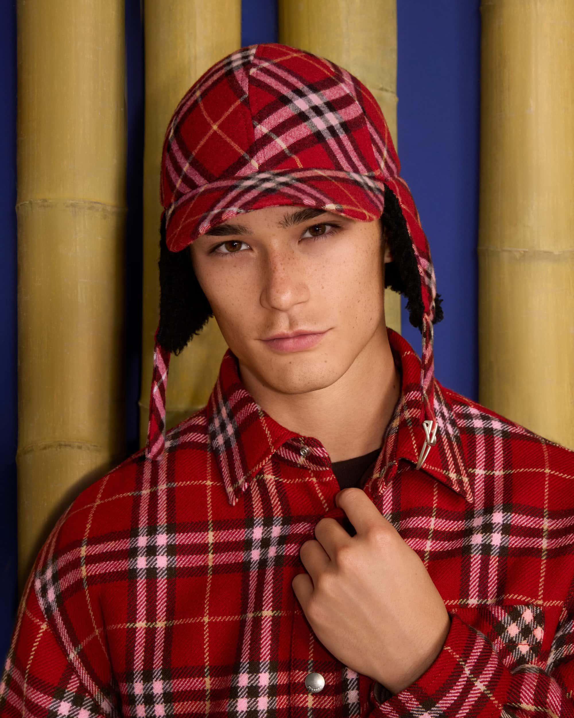 Man wearing a red plaid flapped cap and matching plaid shirt against a bamboo and blue backdrop
