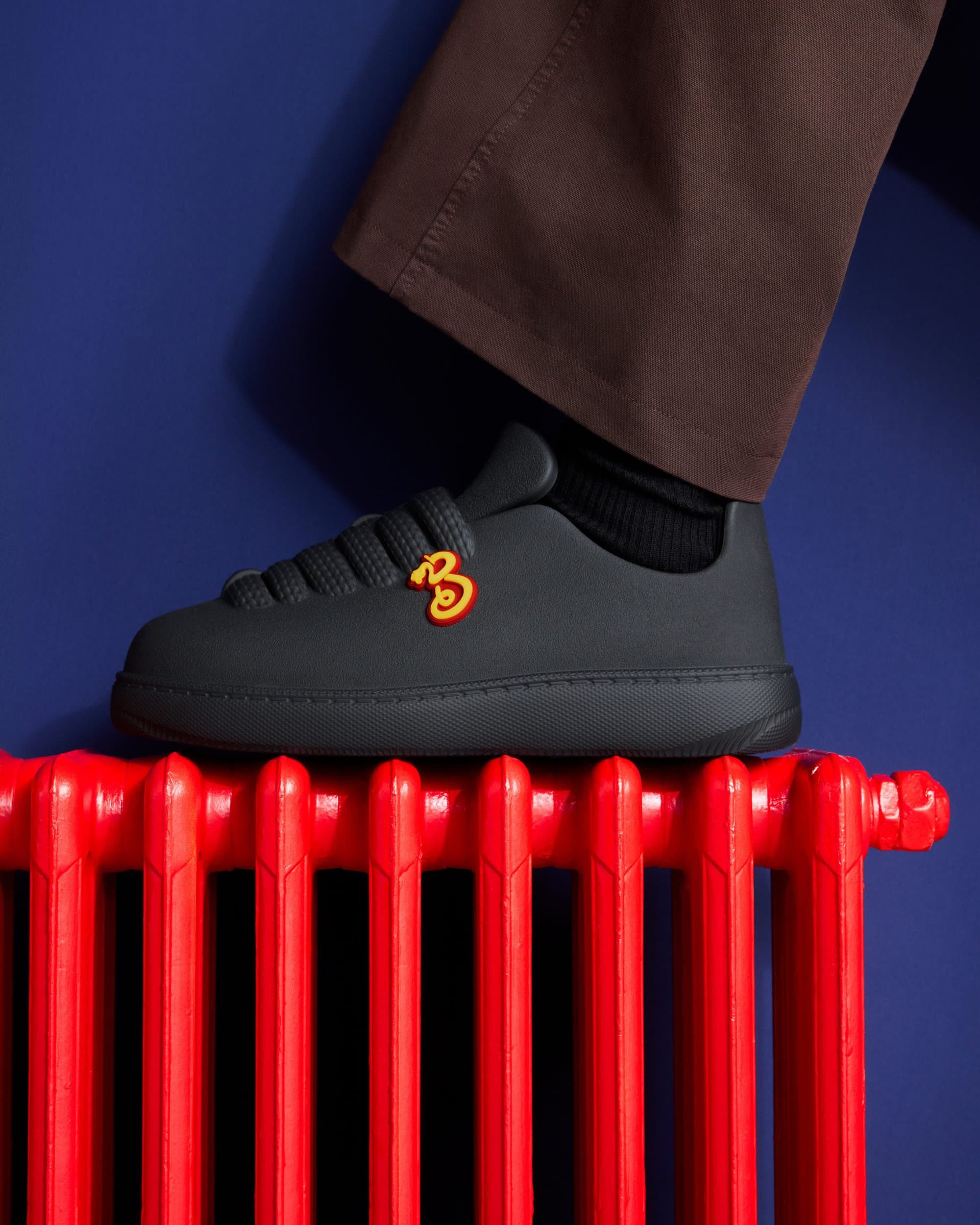 Close-up of a person wearing black sneakers with a red B logo standing on a bright red radiator against a deep blue background.