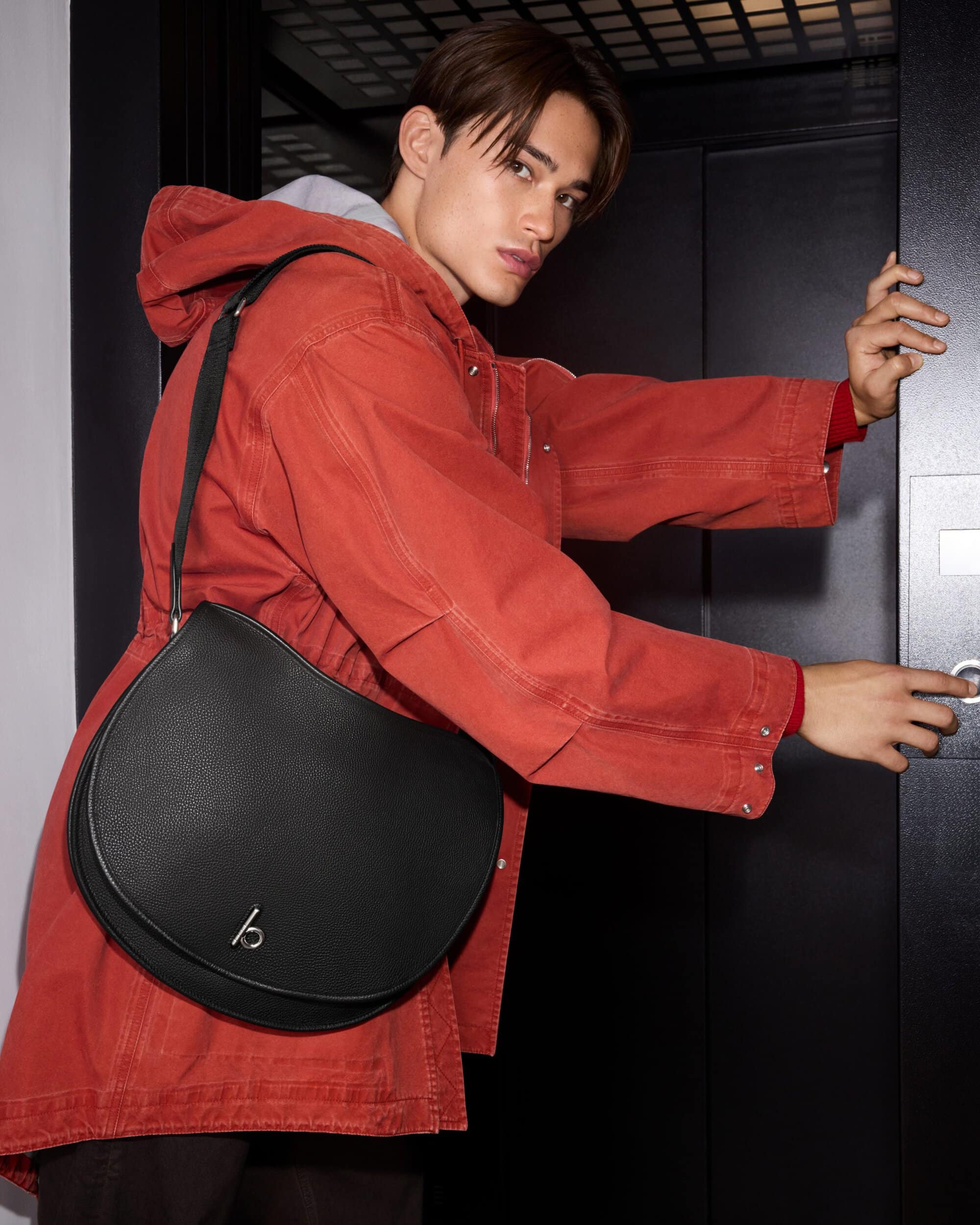 Young man in red jacket wearing a black leather shoulder bag pressing an elevator button.