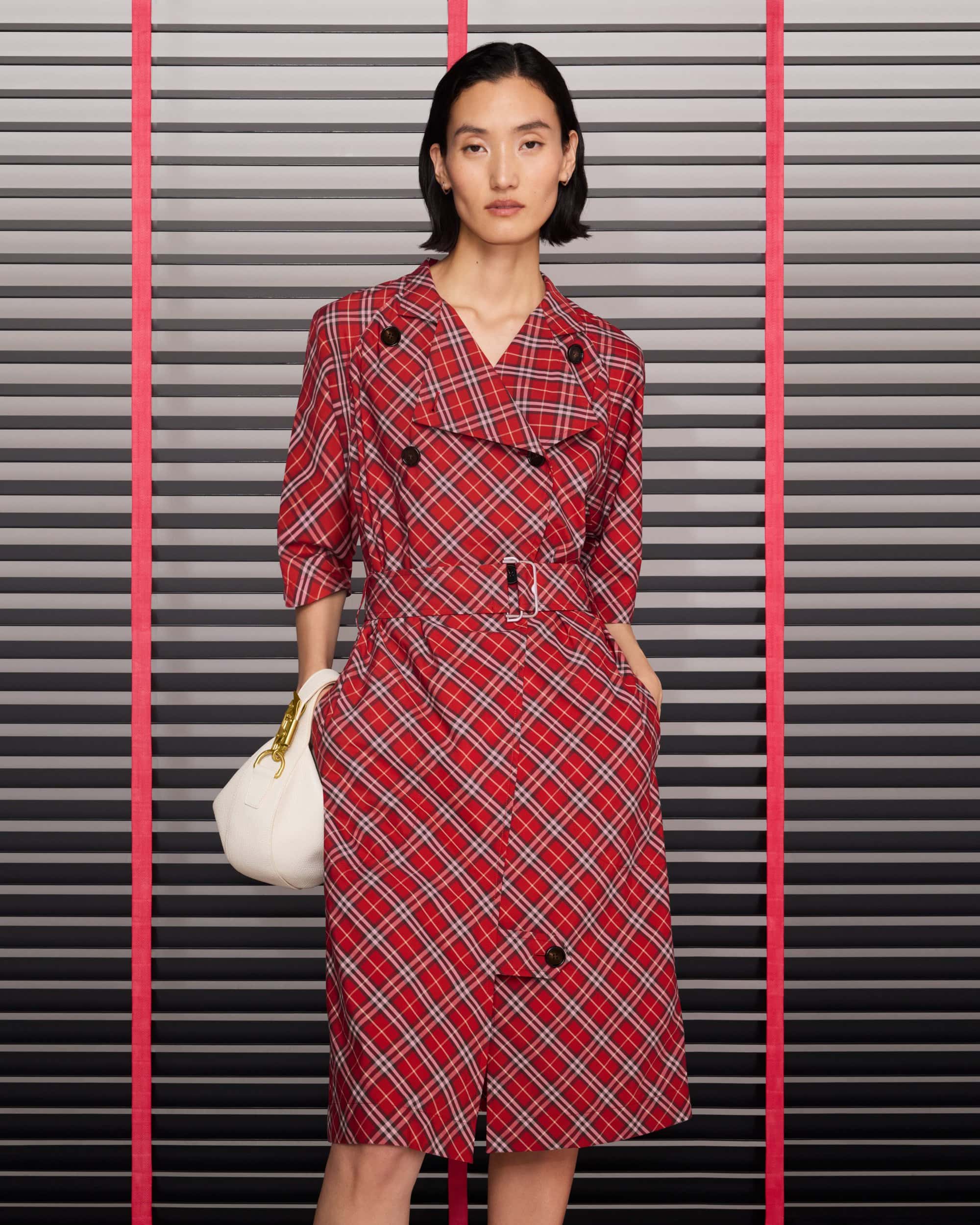 Model wearing a red plaid trench coat and holding a white handbag against a slatted background with pink vertical lines