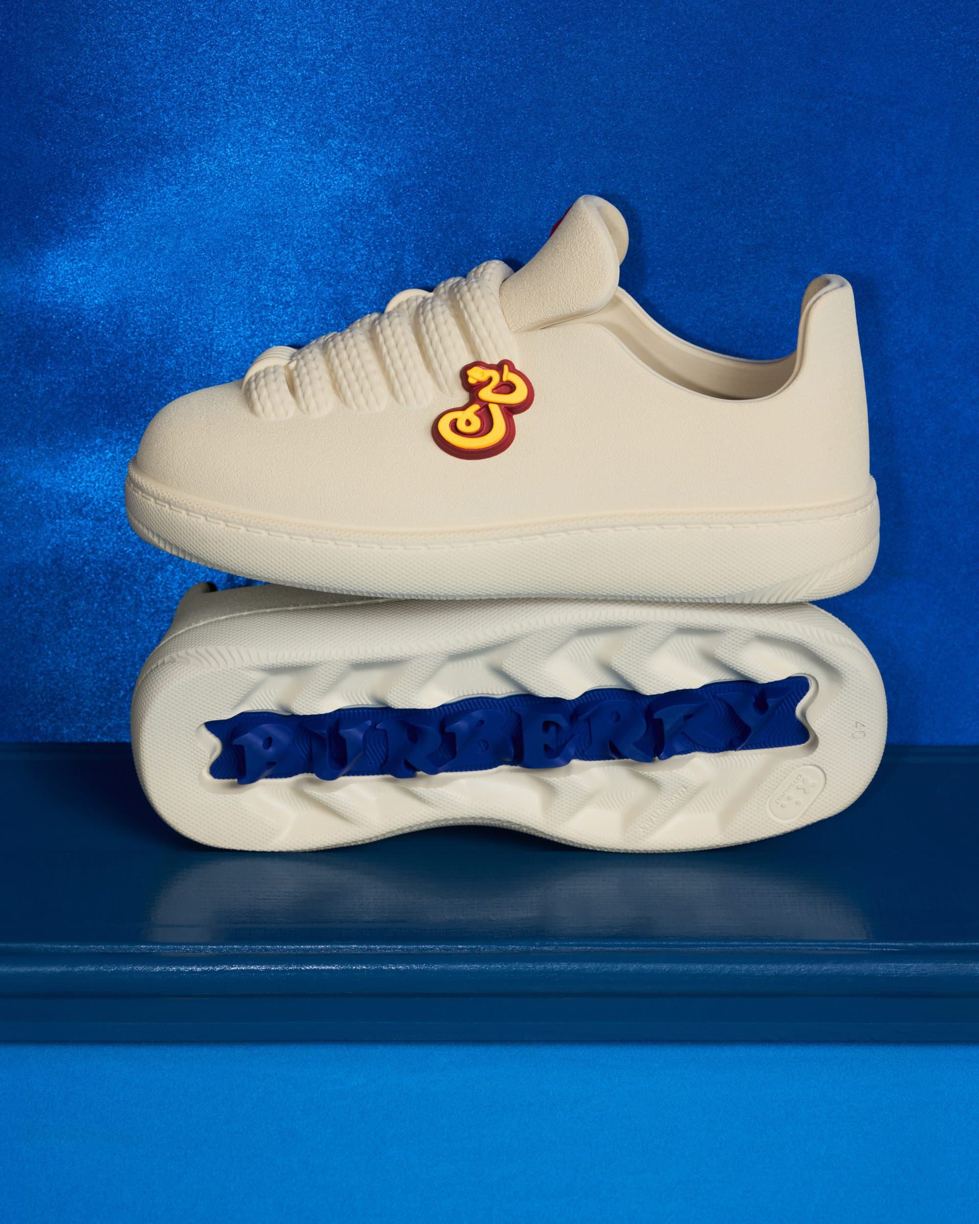Two off-white sneakers stacked vertically against a vibrant blue textured background for Burberry Year of the Snake campaign