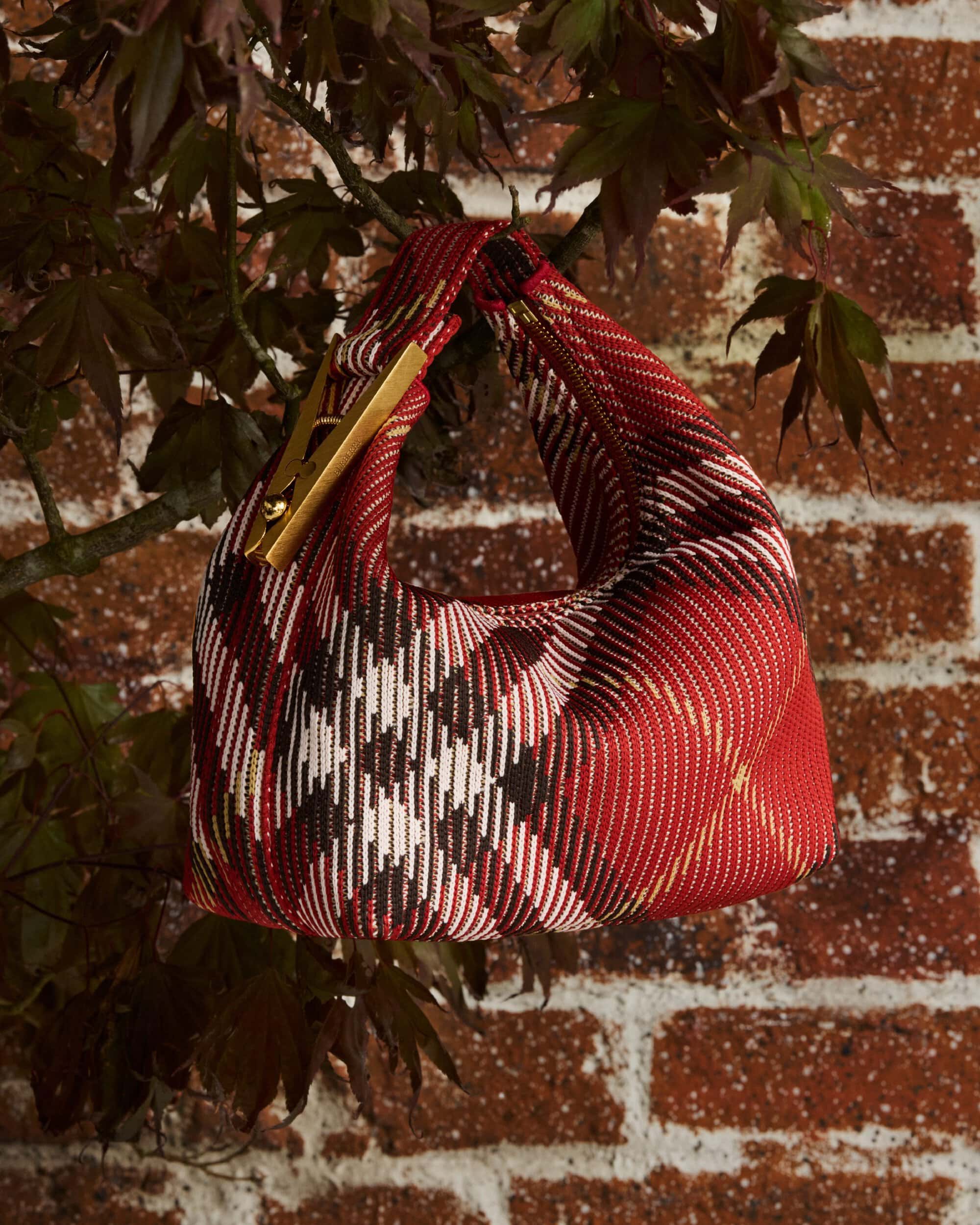 Red plaid handbag hanging from a leafy tree branch against a weathered red brick wall background.