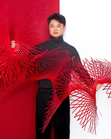 Man holding intricate red woven sculptural structure against split red and white studio background