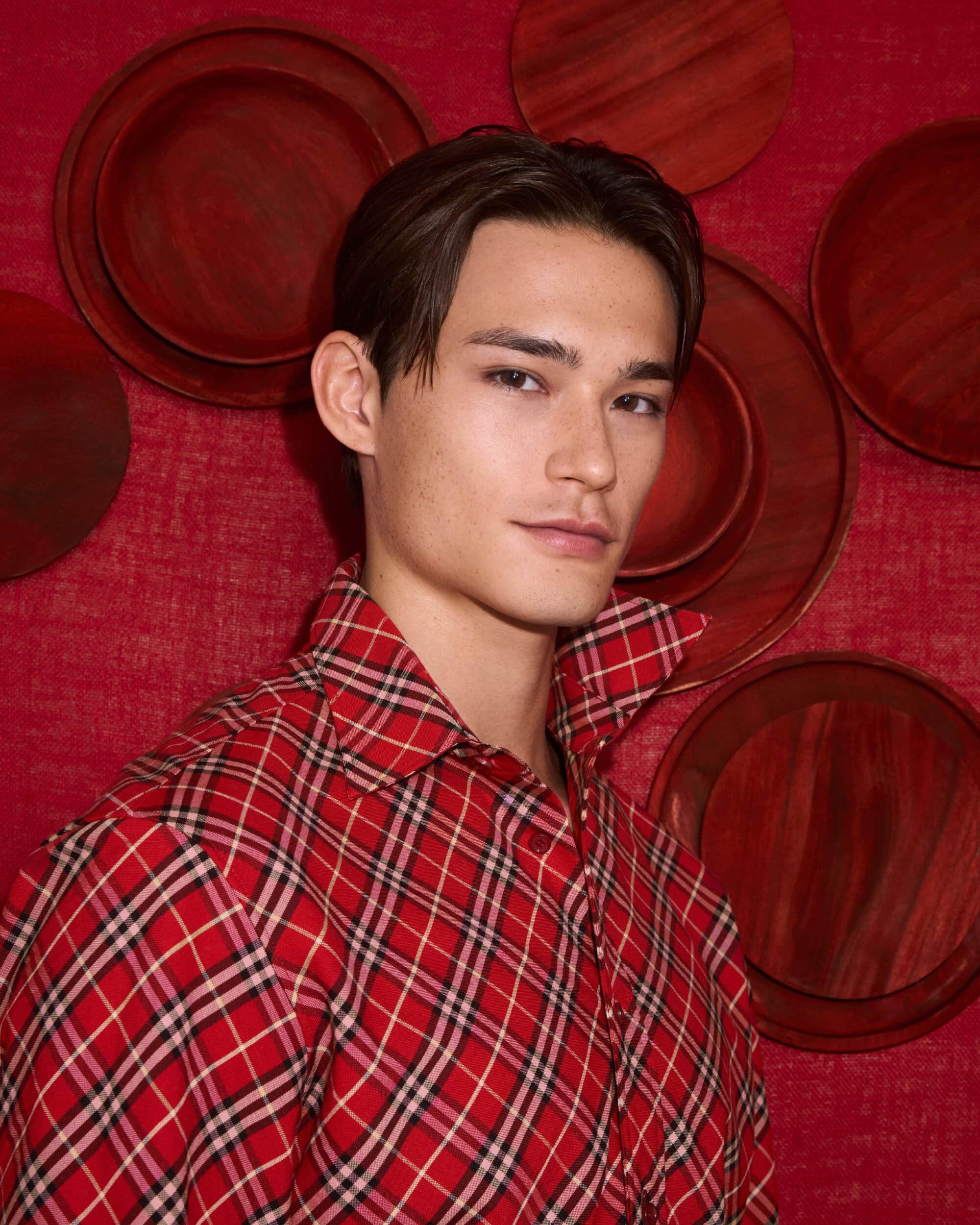 Man wearing a red plaid shirt posing against a textured red wall with decorative round plates.