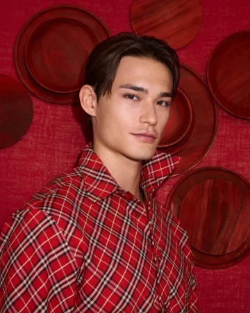 Man wearing a red plaid shirt posing against a textured red wall with decorative round plates.