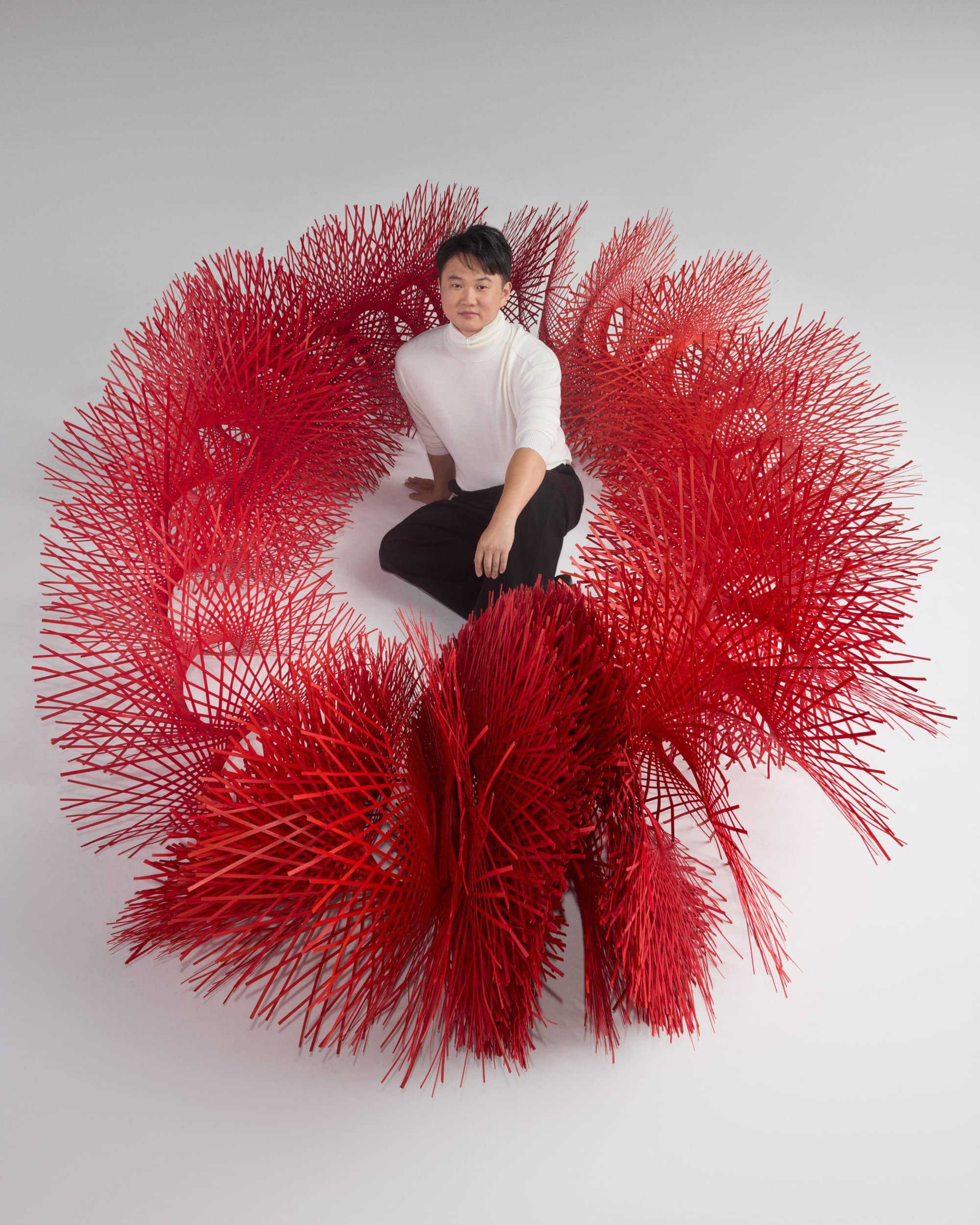 Man kneeling inside a large circular red bamboo sculptural installation against a white background.