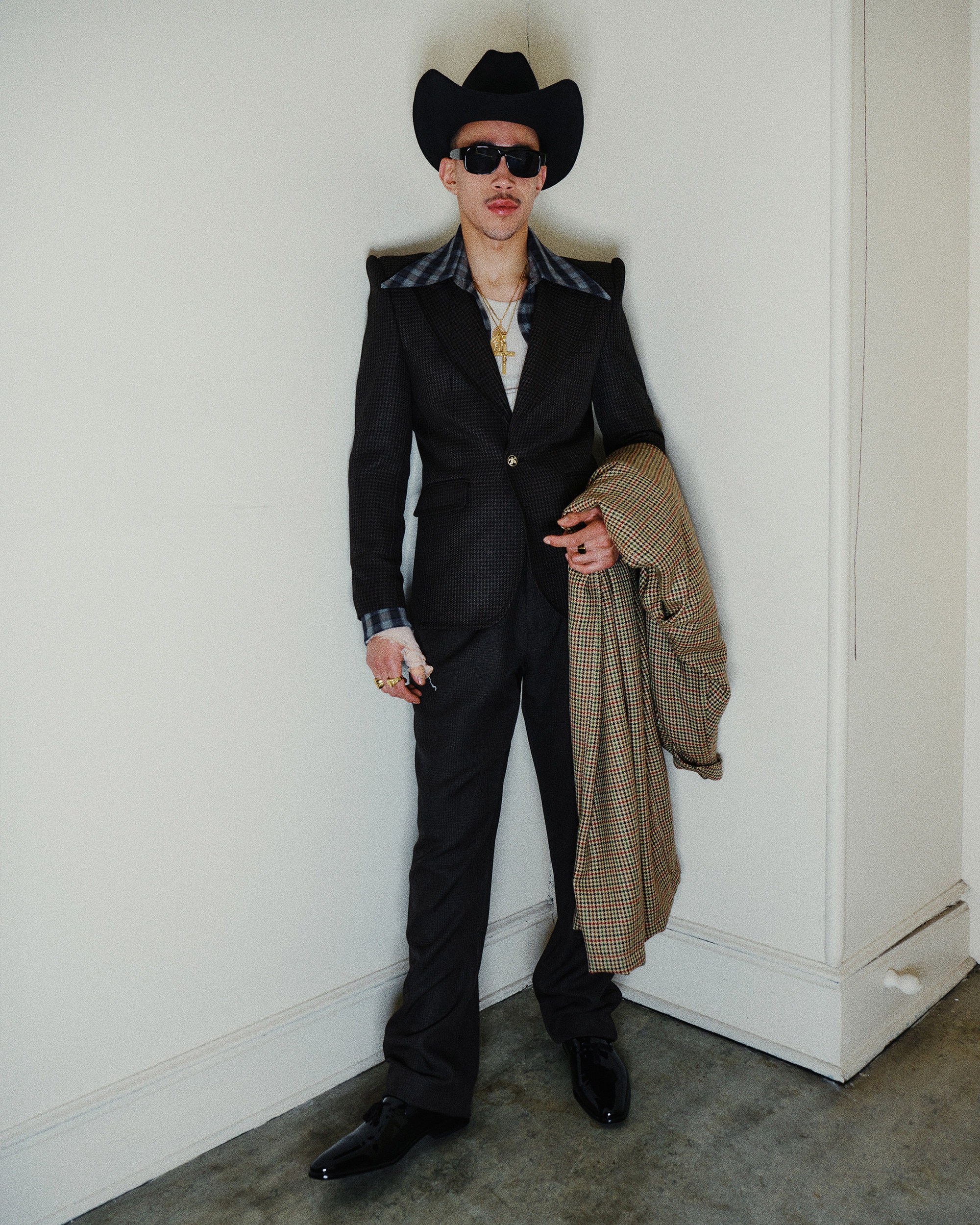 Man wearing a dark suit, cowboy hat, and sunglasses holding a houndstooth coat against an interior wall.