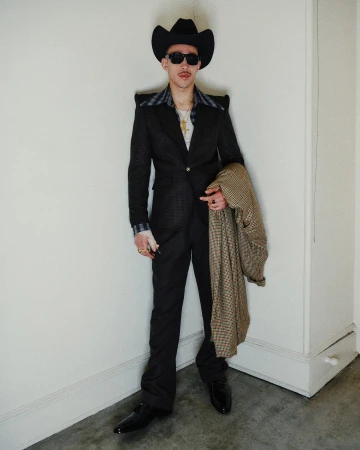 Man wearing a dark suit, cowboy hat, and sunglasses holding a houndstooth coat against an interior wall.