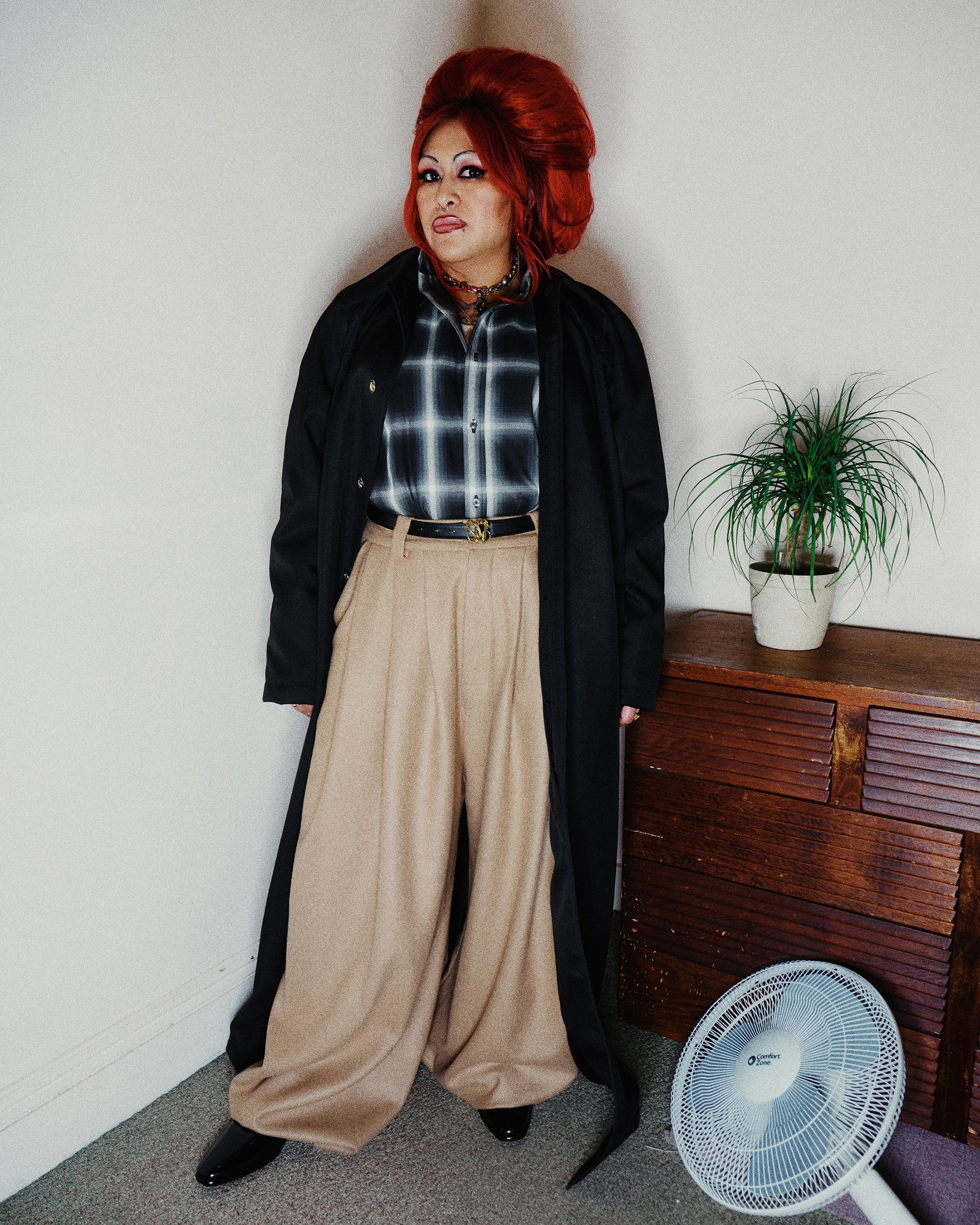 Person with voluminous red hair wearing a black coat, plaid shirt, and beige trousers standing indoors.