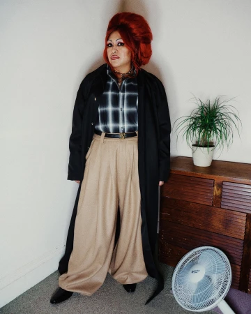 Person with voluminous red hair wearing a black coat, plaid shirt, and beige trousers standing indoors.