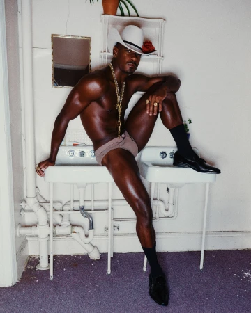 Muscular man wearing a white cowboy hat and briefs seated on a bathroom sink in an ad campaign by Willy Chavarria