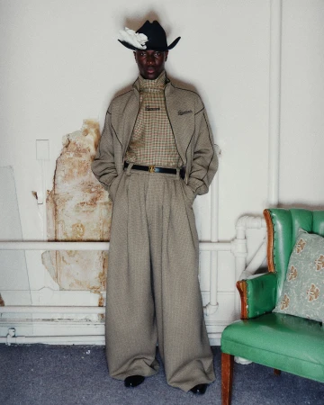 Man wearing a cowboy hat, plaid jacket, and wide-legged trousers standing next to a green armchair.